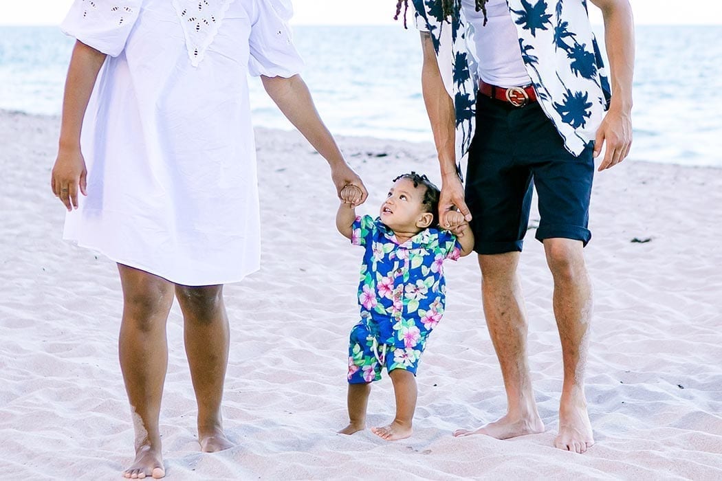 mother and father hold hands with son on beach | son taking first steps on beach | beach family photos in fort lauderdale