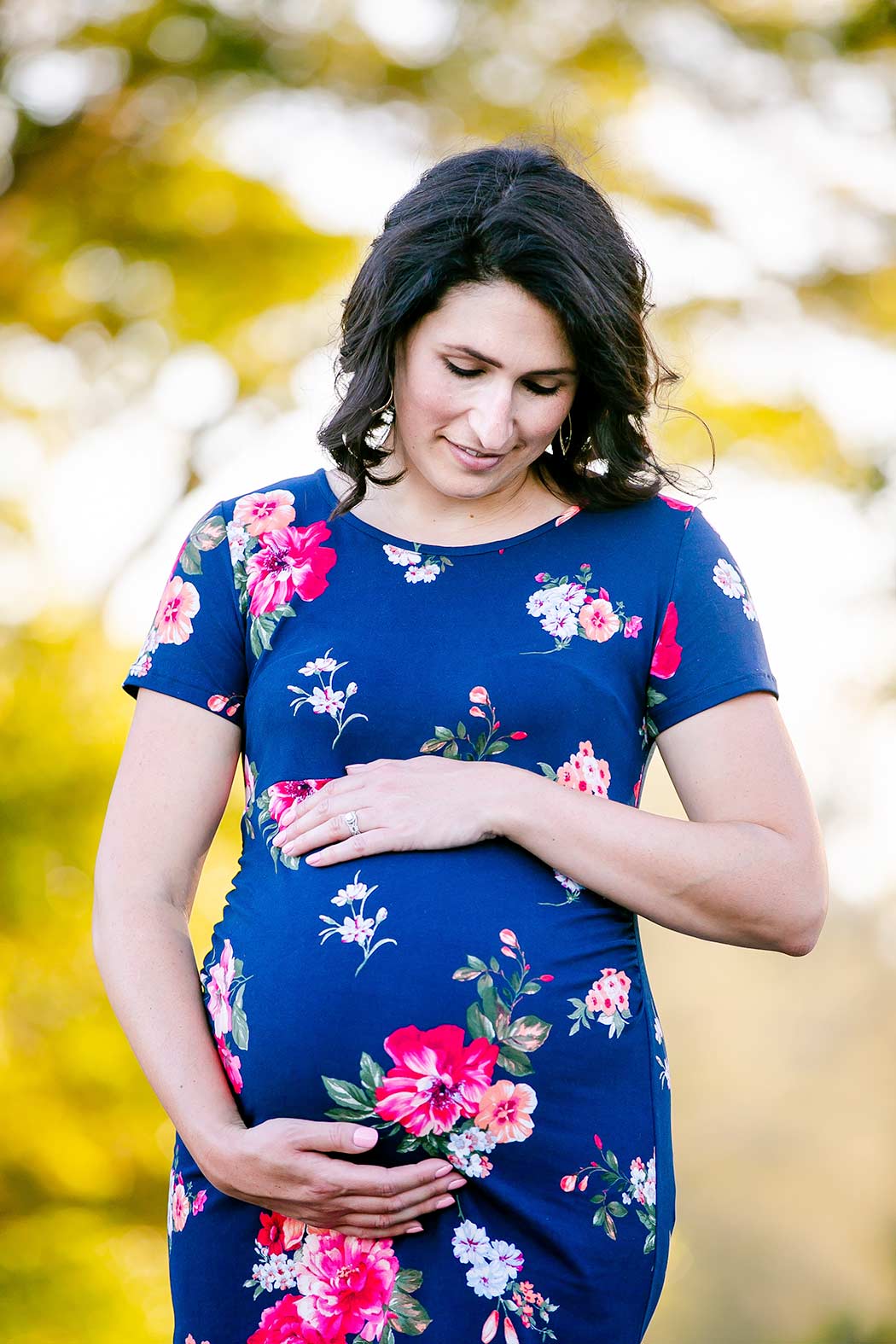 woman holds belly during maternity photoshoot pose | blue floral maternity dress | maternity photoshoot tree tops park fort lauderdale | maternity photographer south florida